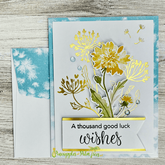 A handmade card featuring a beautiful arrangement of dandelions in shades of yellow and green. The dandelions are accented with gold foil and small water droplets. A banner at the bottom reads "A thousand good luck wishes." The card is paired with a matching blue envelope with a watercolor design.
