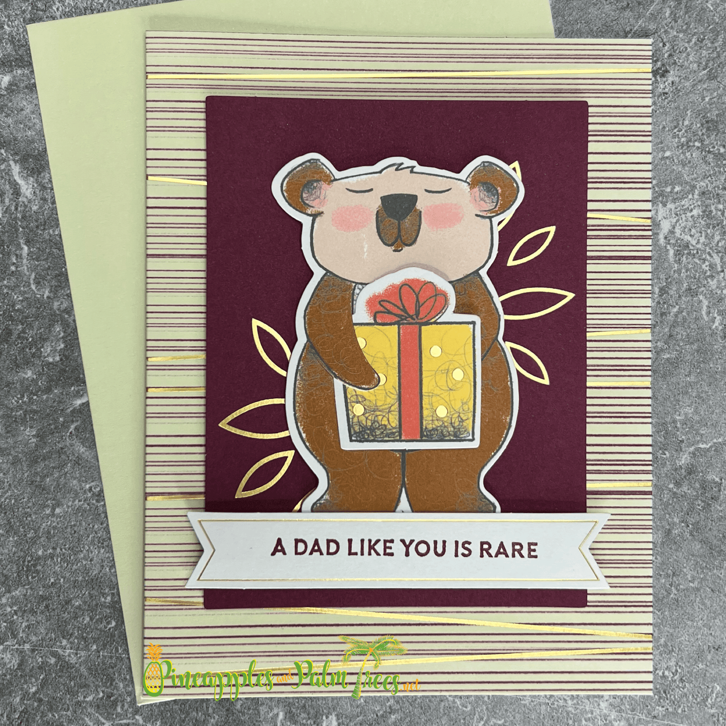 A handmade Father's Day card featuring a cute brown bear holding a brightly wrapped gift. The bear is surrounded by gold foil leaves and stands on a burgundy striped background. The card includes a banner with the sentiment "A Dad Like You is Rare." A matching green envelope is included.