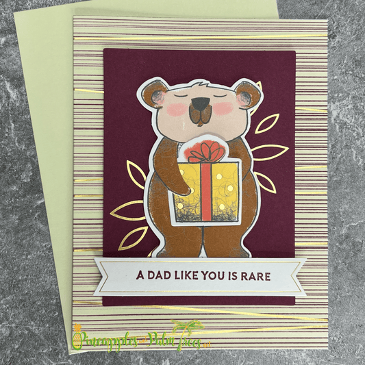 A handmade Father's Day card featuring a cute brown bear holding a brightly wrapped gift. The bear is surrounded by gold foil leaves and stands on a burgundy striped background. The card includes a banner with the sentiment "A Dad Like You is Rare." A matching green envelope is included.
