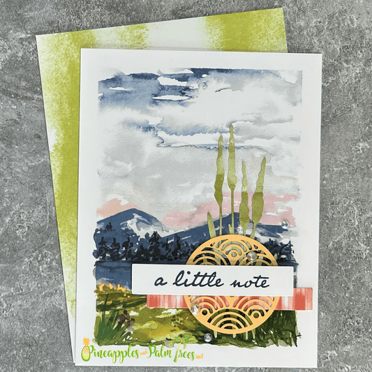 A handmade card with a scenic landscape. The card features a watercolor painting of a mountain range, a sunset, and tall grasses. A gold foil circle with a geometric pattern is layered over the scene, and the sentiment "a little note" is written on a red ribbon. The card is paired with a green envelope.