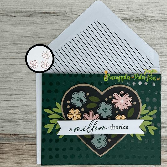A handmade thank-you card featuring a heart-shaped design filled with colorful flowers. The card has a dark green background with a polka-dot pattern. A banner at the bottom reads "a million thanks." A small circle shows a stamp of flowers inside. A matching white envelope with a striped pattern is included.