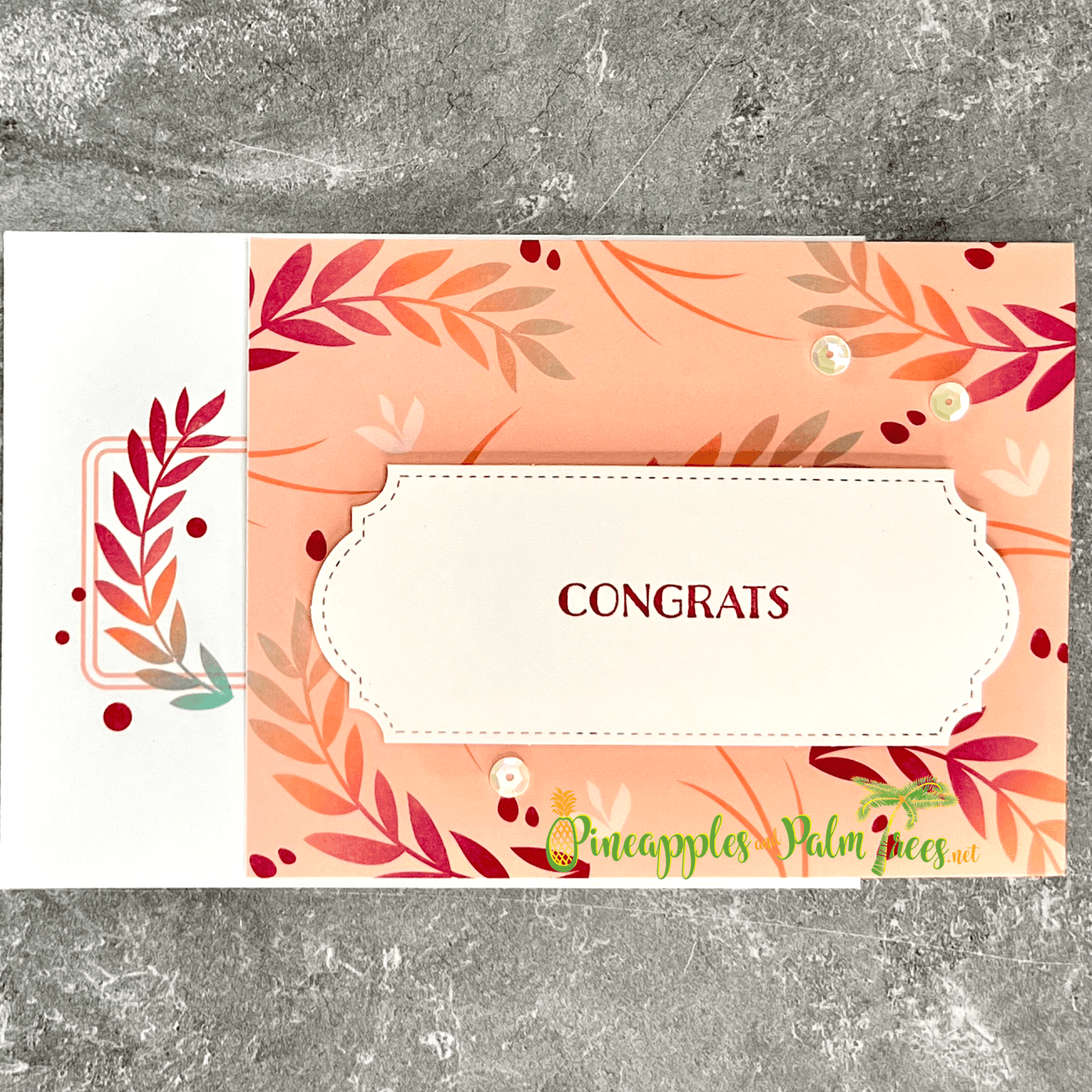 A handmade congratulations card with a fall theme. The card features a peach-colored background with a pattern of autumn leaves. A banner with the word "CONGRATS" is layered over the leaves. The card is finished with a few sequins and a matching white envelope with coordinating pattern.