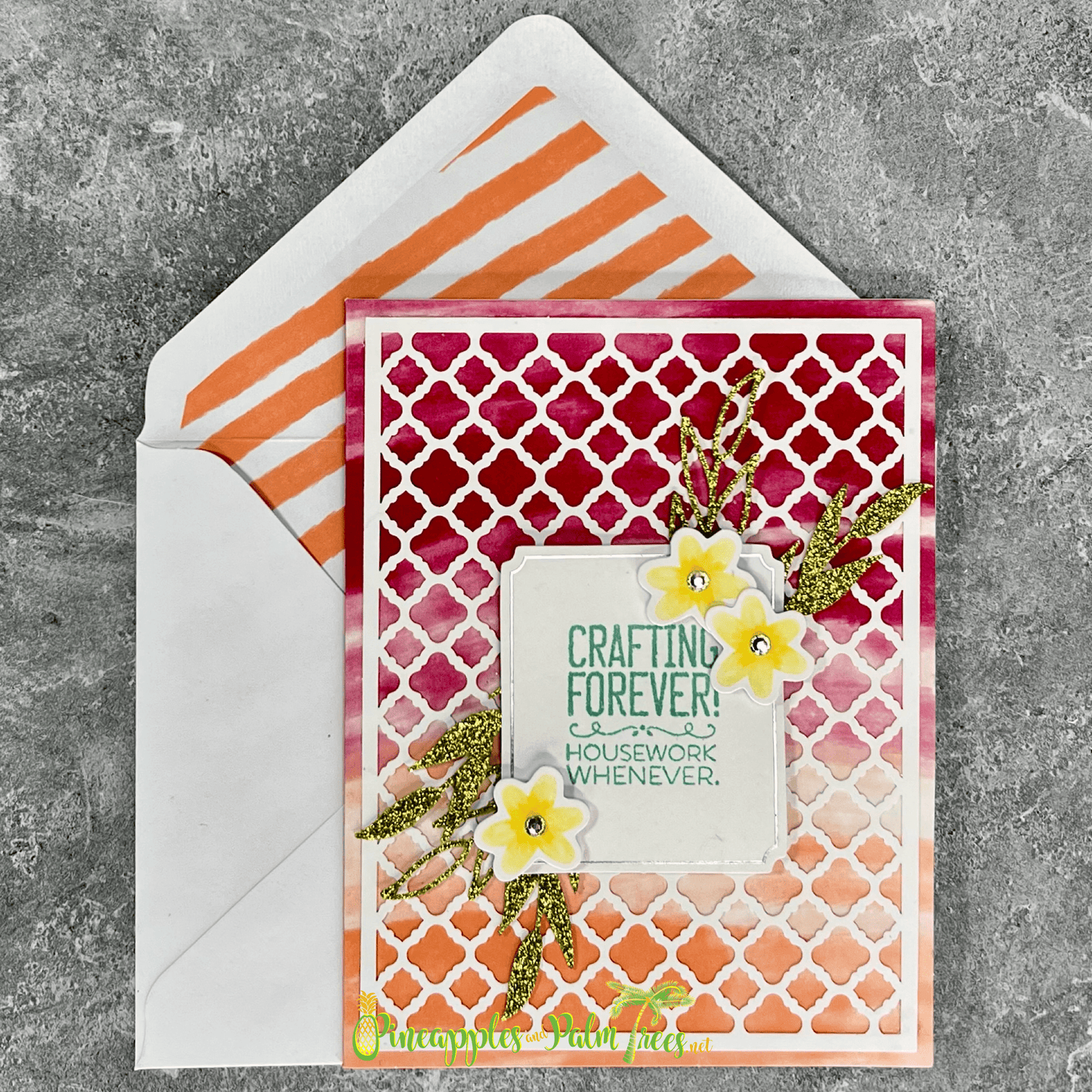 A handmade card with a watercolor background in shades of pink and orange. The card features a square panel with a trellis pattern and the sentiment "CRAFTING FOREVER! HOUSEWORK WHENEVER." Small flowers and green glitter leaves are added to the panel. The card is paired with a white envelope with a striped pattern inside.