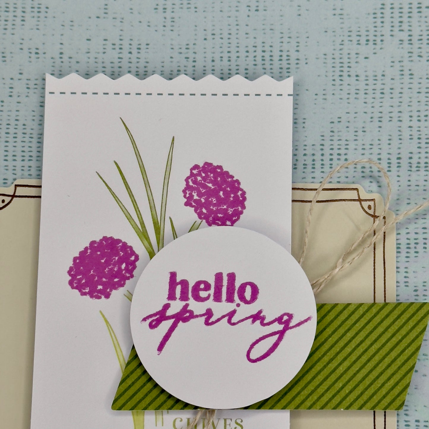 Close up of the greeting card with the words ‘Hello Spring’ surrounded by green chive stems and simple floral accents, shown with its matching envelope — light, fresh, and handcrafted.