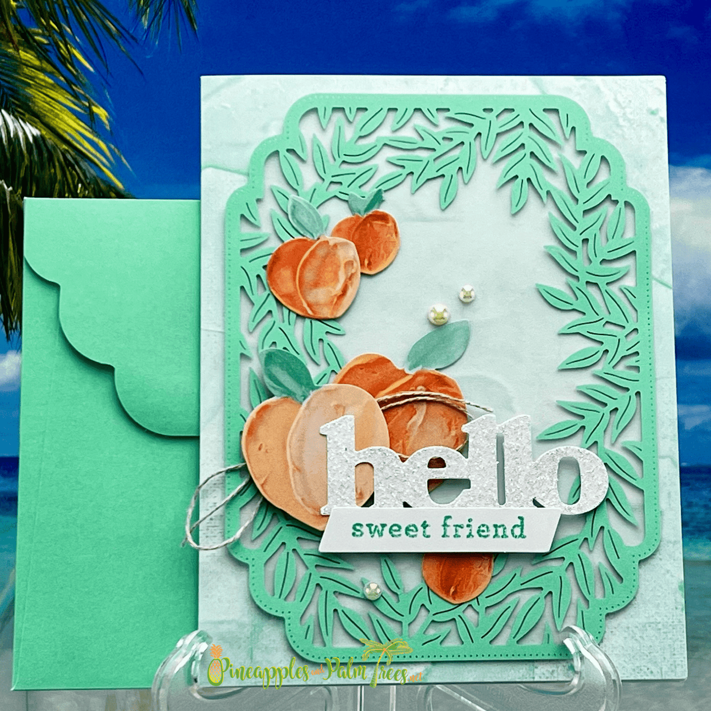The greeting card is handmade card with a peach theme. The card is primarily white with a watercolor wash of peach and green. A die-cut frame surrounds the main design, which features several watercolor peaches, a banner with the words "hello sweet friend" in silver glitter, and a few small white pearls. A coordinating green envelope is paired with the card.