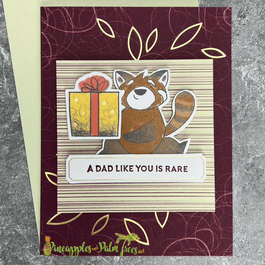 A handmade Father's Day card featuring a cute red panda holding a brightly wrapped gift. The panda is surrounded by gold foil leaves and stands on a burgundy striped background. The card includes a banner with the sentiment "A Dad Like You is Rare." A matching green envelope is included.