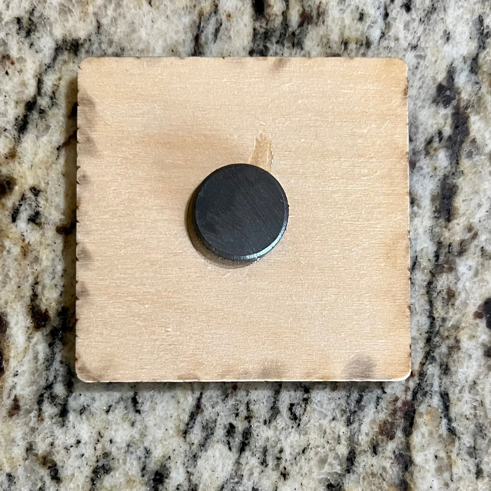 Backside of a 2”x2” wooden square with a magnet in the center.  Background is tan/brown granite.