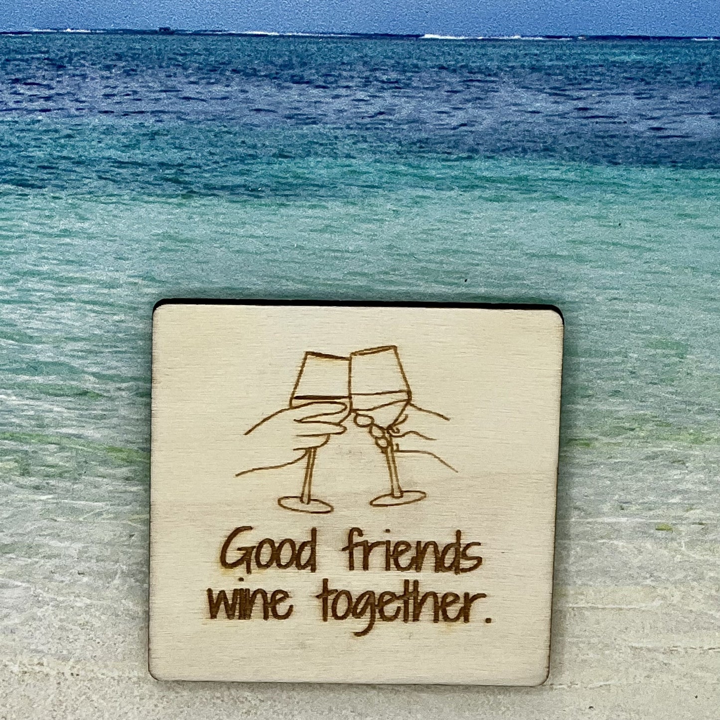 2”x2” wooden square with “Good Friends Wine Together“ and toasting with wine, laser engraved. Background scene is a tropical ocean.