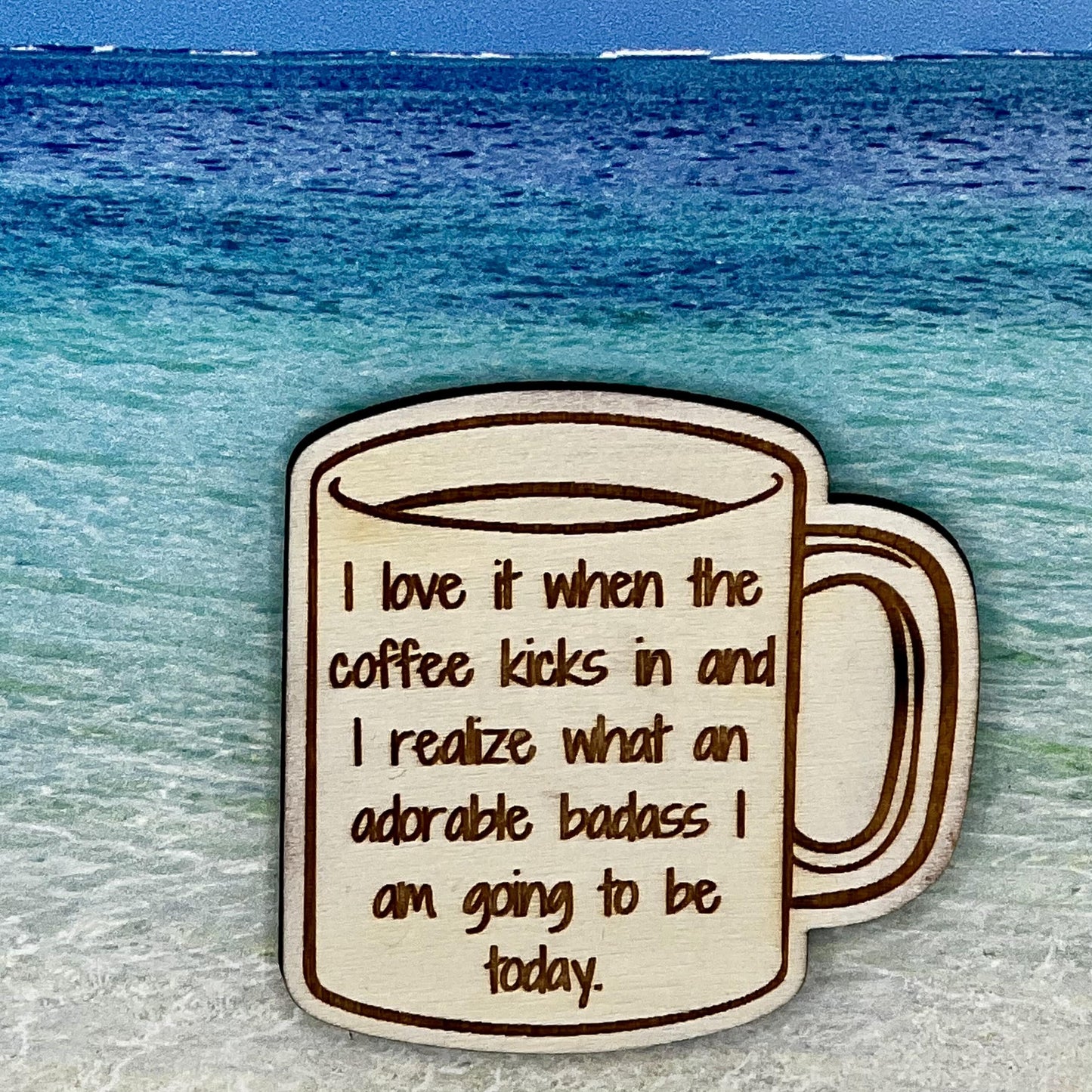 Wood outline of a coffee mug with “ I Love it When the Coffee Kicks In and I Realize What an Adorable Badass I Am Going to be Today.“ laser engraved. Background scene is a tropical ocean.