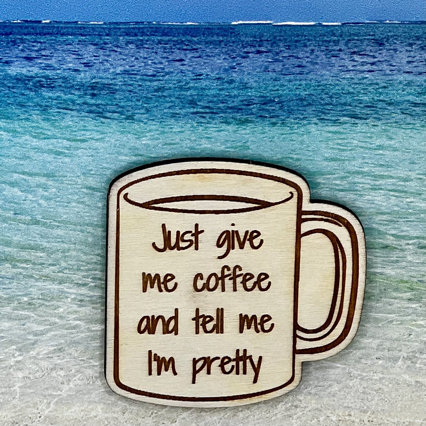 Wood outline of a coffee mug with “Just Give Me Coffee and Tell Me I'm Pretty“ laser engraved. Background scene is a tropical ocean.