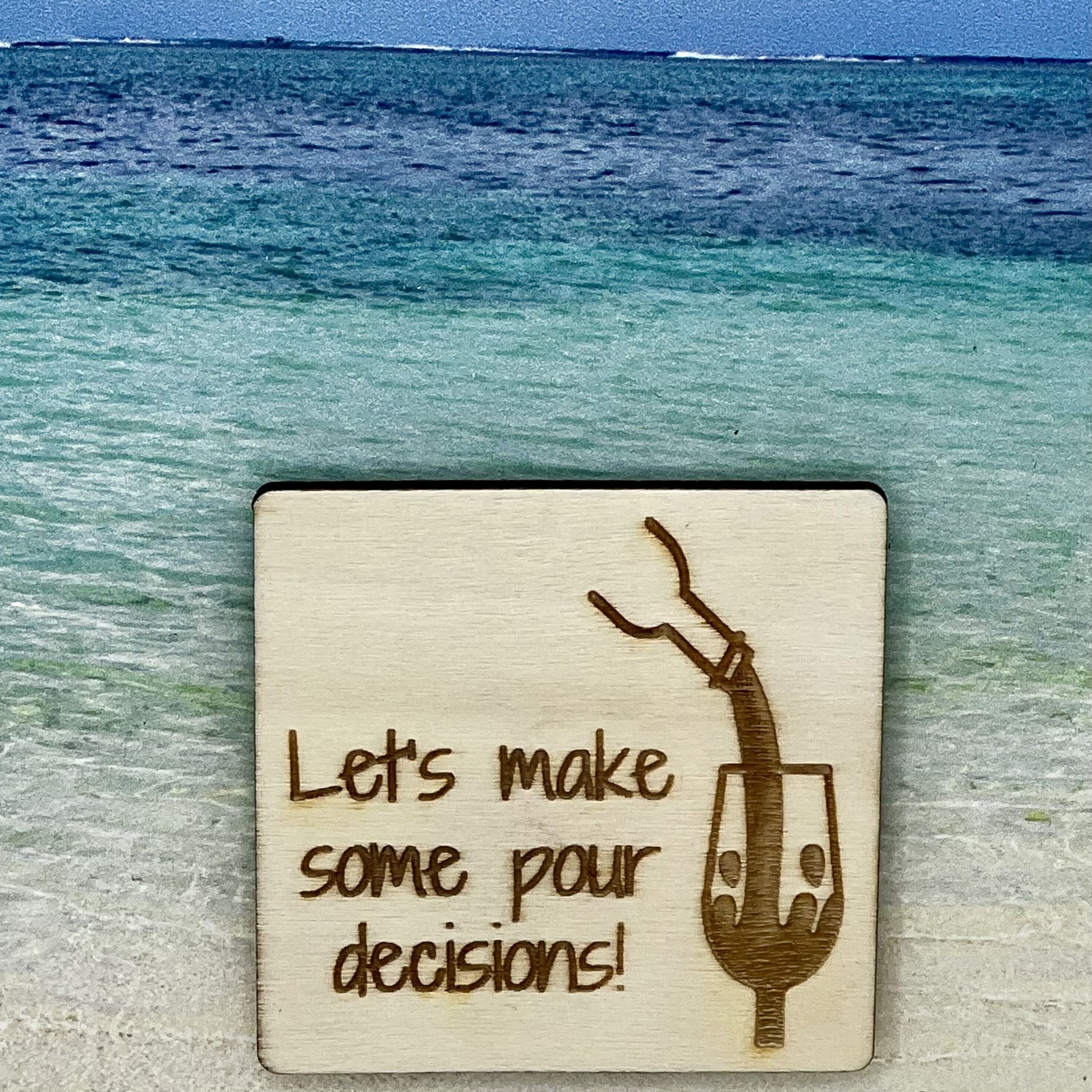 2”x2” wooden square with “ Let's Make Some Pour Decisions!“ and wine being poured into a wine glass, laser engraved. Background scene is a tropical ocean.