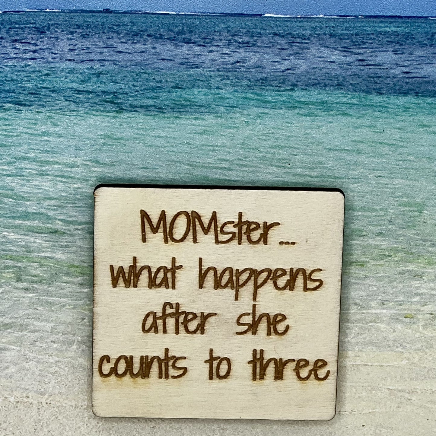 2”x2” wooden square with “MOMster... What Happens After She Counts to Three“ laser engraved. Background scene is a tropical ocean.