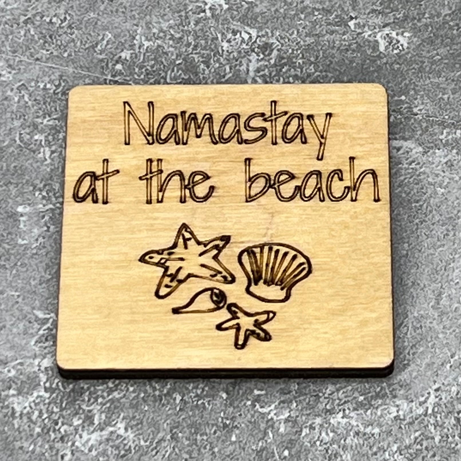 2”x2” wooden square with “Namastay at the Beach“ and some miscellaneous sea shells laser engraved. Background is gray.