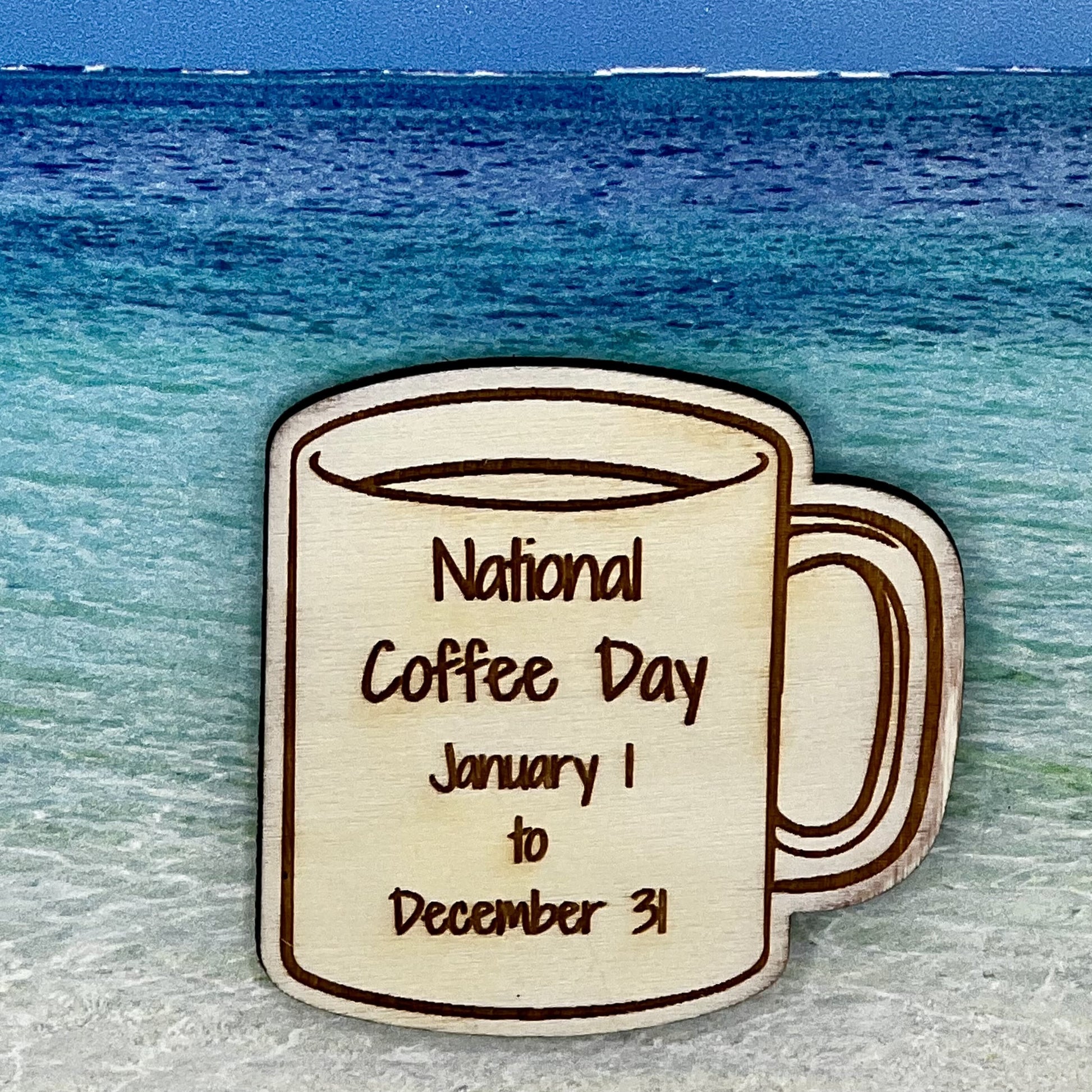 Wood outline of a coffee mug with “National Coffee Day January 1 to December 31“ laser engraved. Background scene is a tropical ocean.