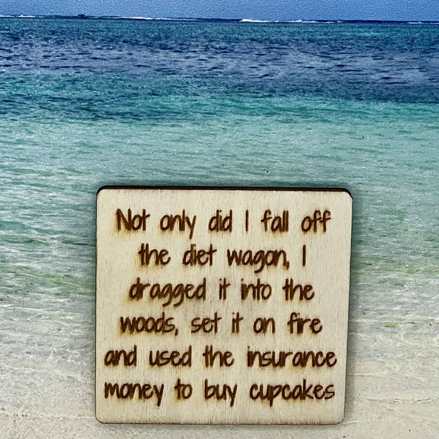 2”x2” wooden square with “ Not Only Did I Fall Off the Diet Wagon, I Dragged It into the Woods, Set It on Fire and Used the Insurance Money to Buy Cupcakes“ laser engraved. Background scene is a tropical ocean.