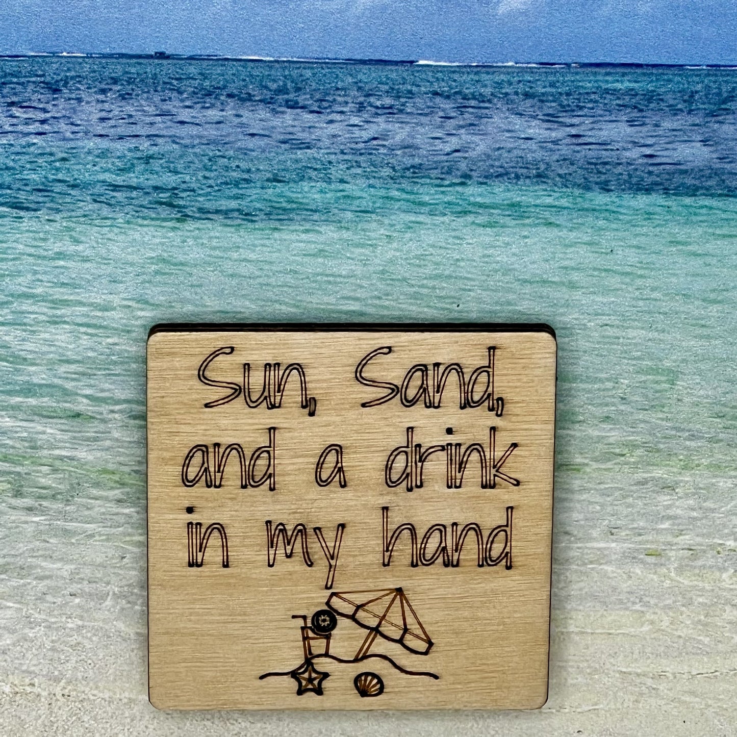 2”x2” wooden square with “Sun, Sand, and a Drink in My Hand" and a beach scene, laser engraved. Background scene is a tropical ocean.