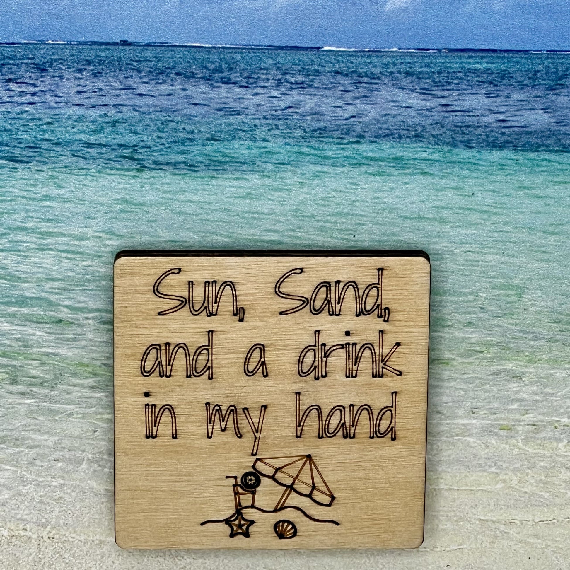 2”x2” wooden square with “Sun, Sand, and a Drink in My Hand" and a beach scene, laser engraved. Background scene is a tropical ocean.