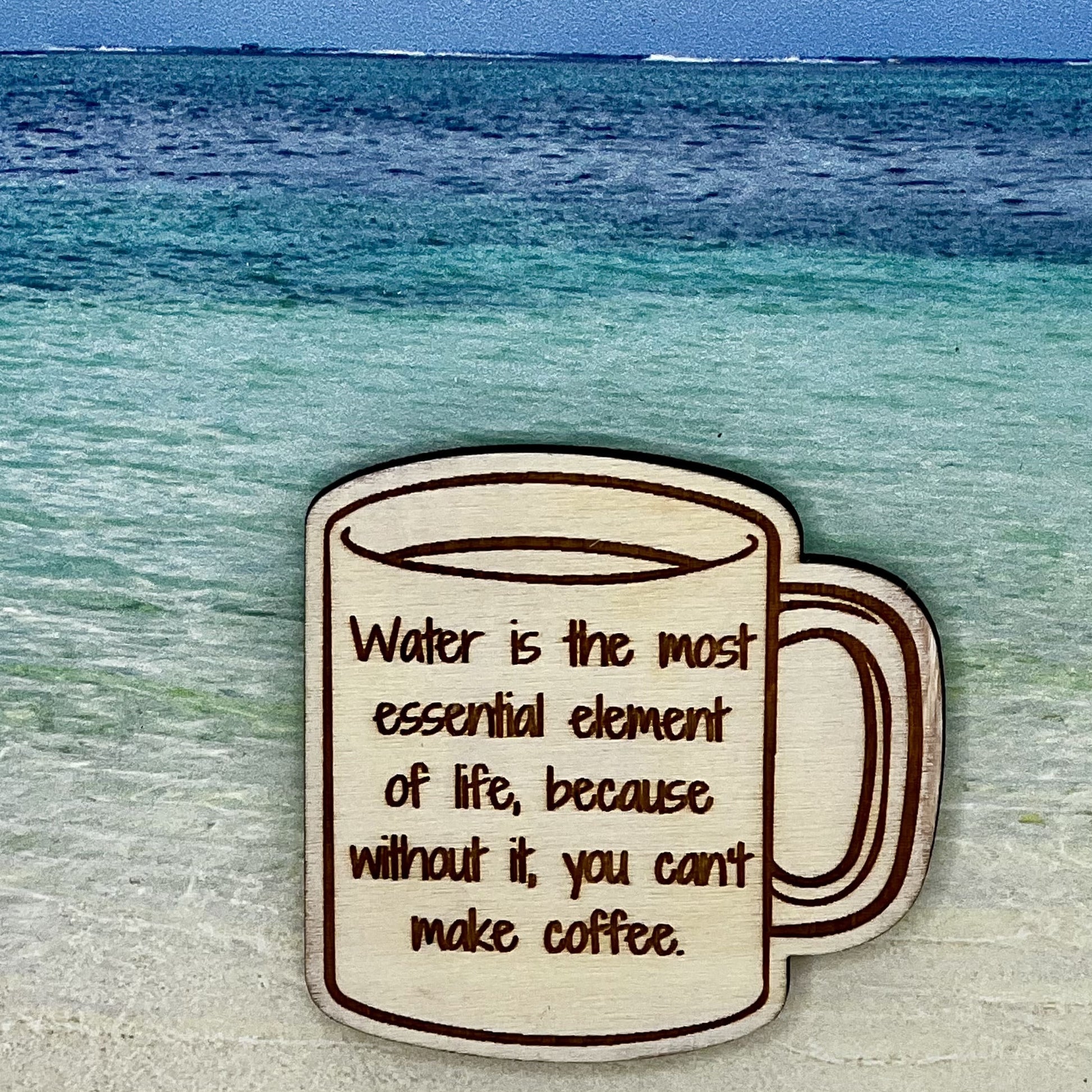 Wood outline of a coffee mug with “Water is the Most Essential Element of Life, Because Without It, You Can't Make Coffee“ laser engraved. Background scene is a tropical ocean.