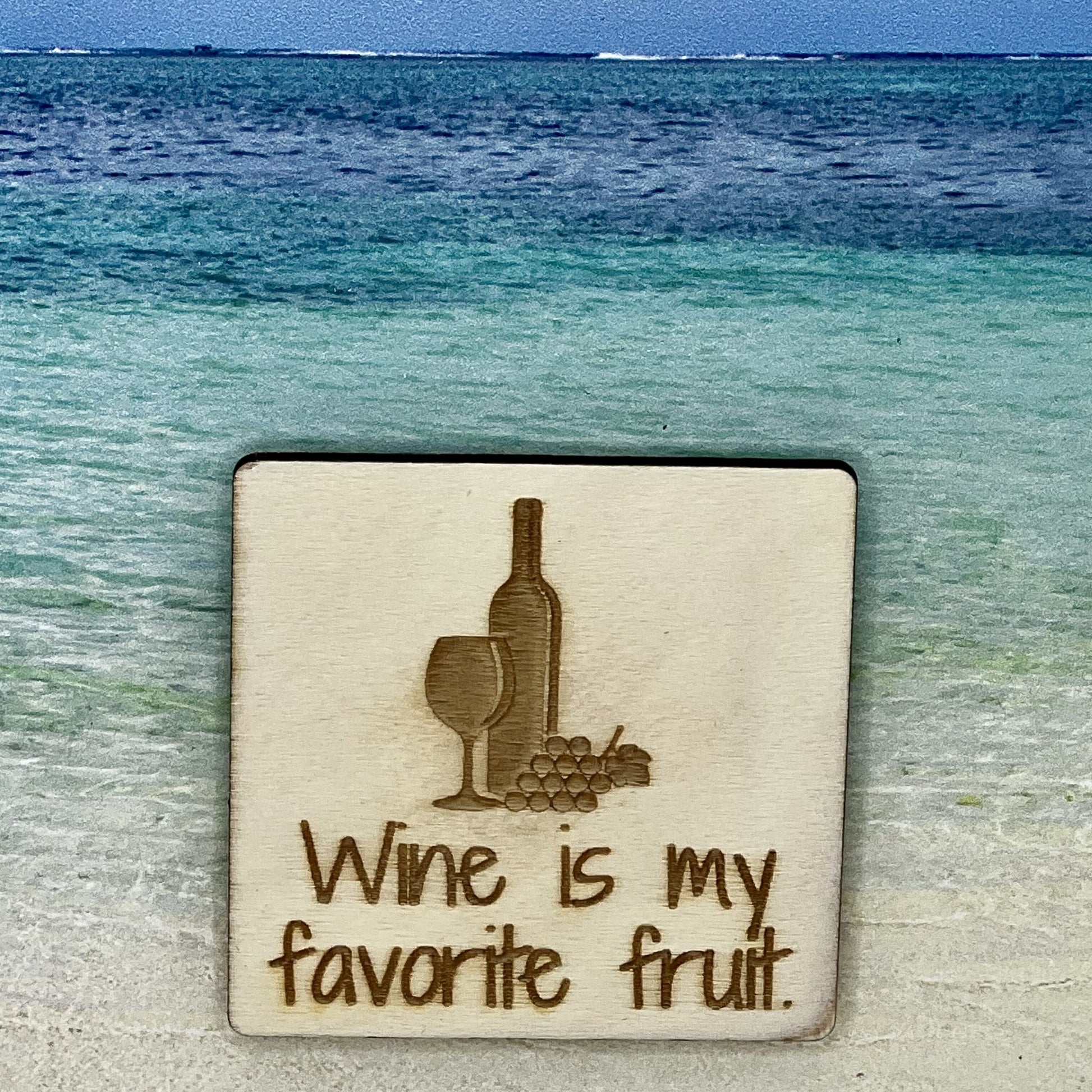 2”x2” wooden square with “Wine is my Favorite Fruit“ and a bottle of wine, wine glass and grapes, laser engraved. Background scene is a tropical ocean.