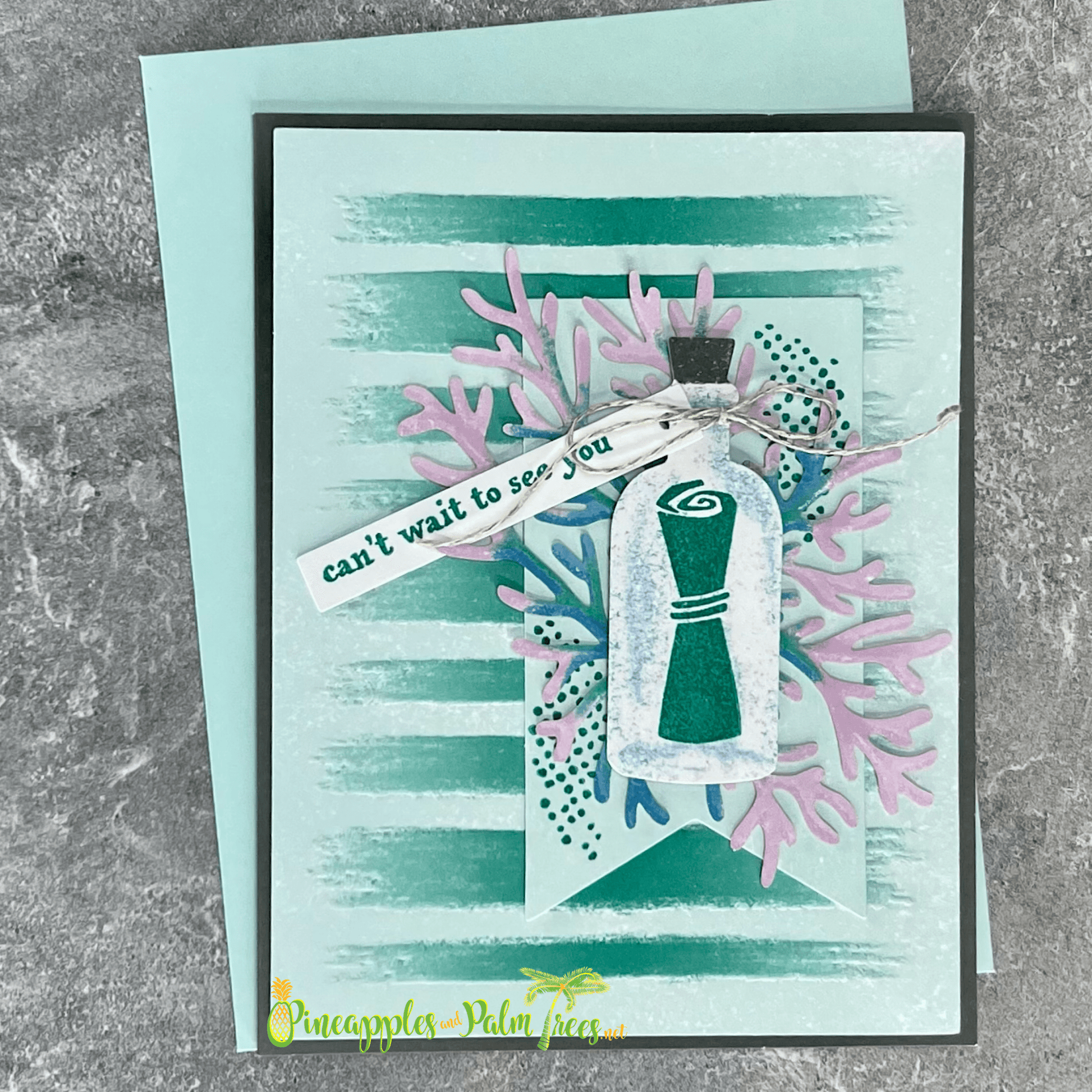 A handmade card with an ocean theme. The card features a message in a bottle, coral, and a banner that reads "can't wait to see you." The background is a watercolor wash in shades of blue and green with stripes. A matching teal envelope is included.