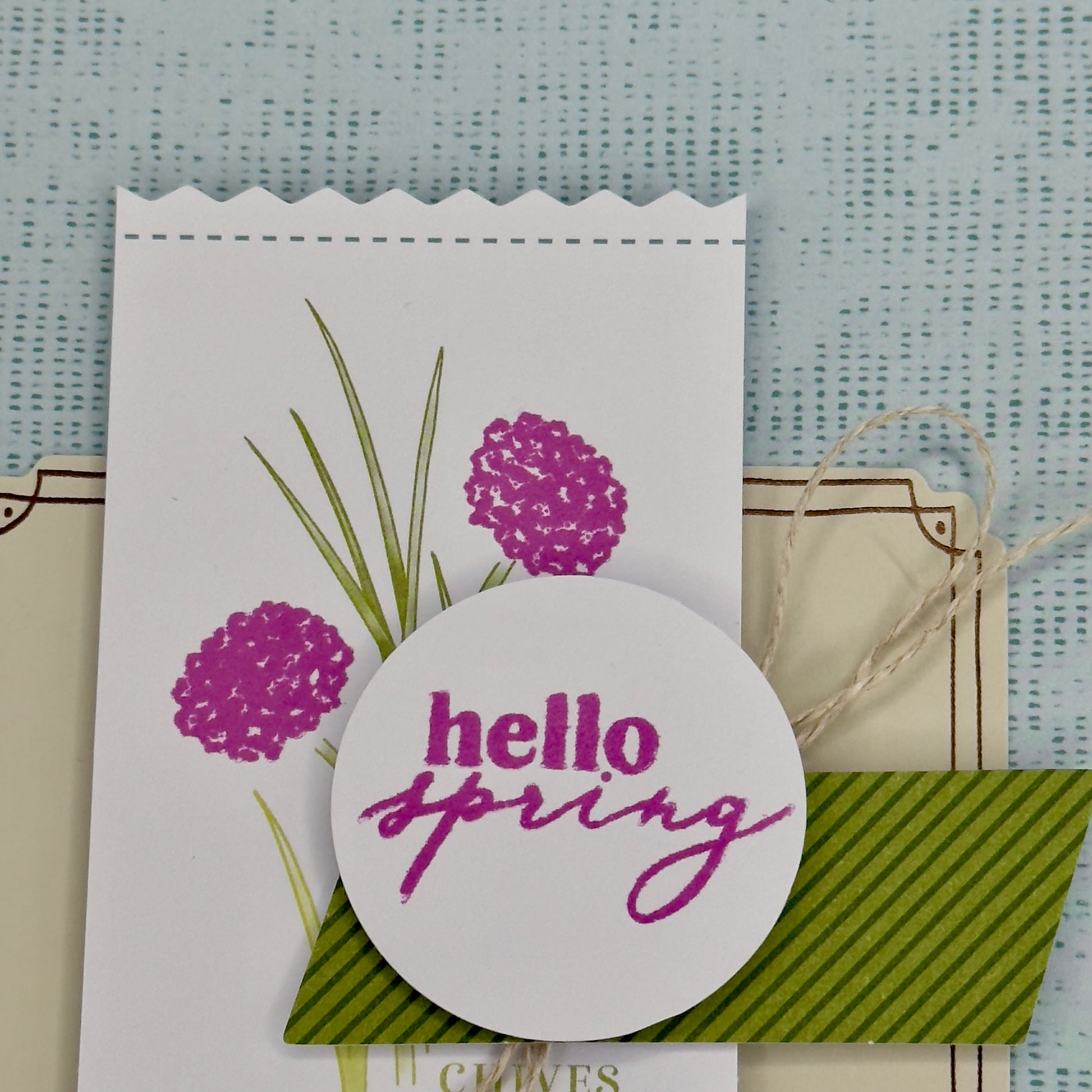 Close up of the greeting card with the words ‘Hello Spring’ surrounded by green chive stems and simple floral accents, shown with its matching envelope — light, fresh, and handcrafted.

