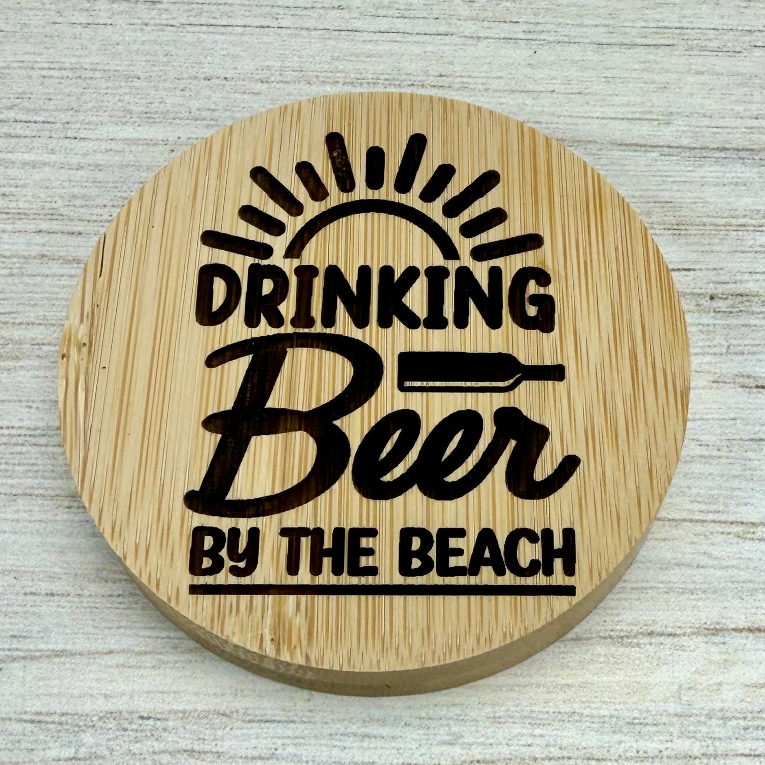 Round laser‑engraved bottle opener with “Drinking Beer by the Beach” design, perfect for summer or beach use.